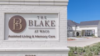 Sign on a white brick wall displaying the logo and name 'The Blake at Waco' with the text 'Assisted Living & Memory Care'. In the background, there is a building with a light-colored exterior and a driveway.