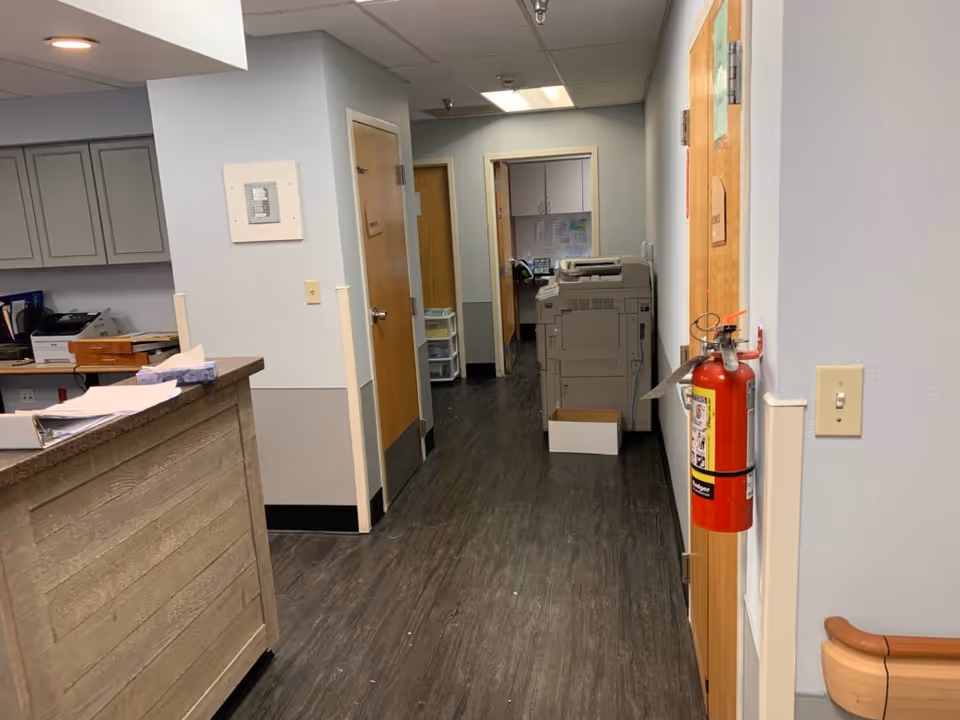 Interior hallway of a facility with a wooden reception desk on the left, closed wooden doors along the hallway, a fire extinguisher mounted on the right wall, and a large printer/copier machine at the end of the hallway.