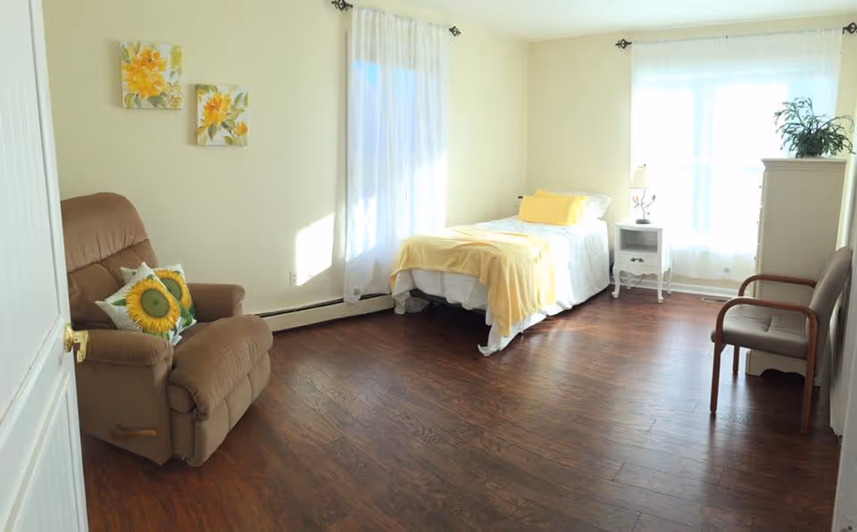 Sunlit assisted living bedroom with a single bed, recliner, nightstand and chairs on a wood floor.