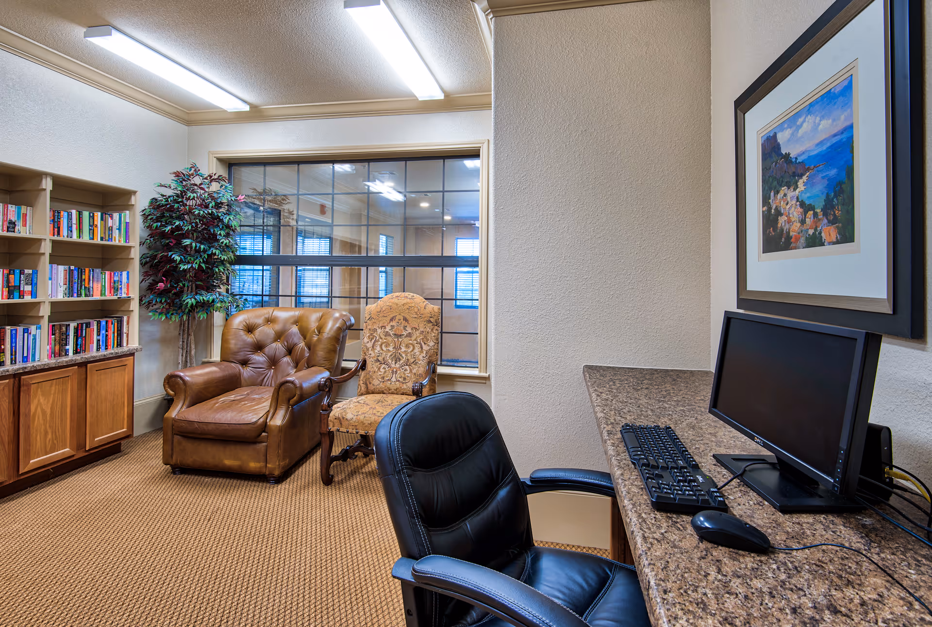 A cozy interior room with a computer workstation featuring a monitor, keyboard, and mouse on a granite countertop. There is a black office chair in front of the desk. In the background, there are two armchairs, one leather and one upholstered, next to a tall artificial plant and a bookshelf filled with books. A framed painting of a coastal scene hangs on the wall above the desk.
