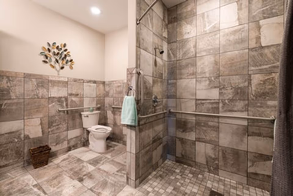 A spacious bathroom with beige and brown tiled walls and floor. The room features a toilet with grab bars on both sides, a walk-in shower with a handheld showerhead and multiple grab bars, a towel hanging on a ring near the shower, and a decorative metal wall art shaped like a tree above the toilet. There is also a small wastebasket on the floor near the toilet.