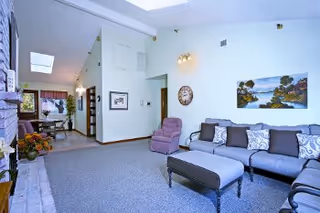 A spacious living room with a gray sectional sofa adorned with patterned pillows, a matching ottoman, and a purple recliner chair. The room features a light gray carpet, a wall clock, and a landscape painting on the wall. In the background, there is a dining area with a table and chairs, and large windows allowing natural light to enter.