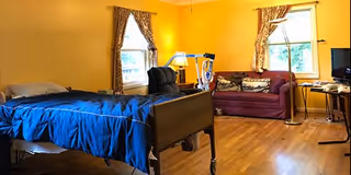 A bright room with yellow walls featuring a hospital-style bed with blue bedding, a black reclining chair, a maroon sofa with patterned cushions, two windows with curtains, a floor lamp, and a television on a stand.