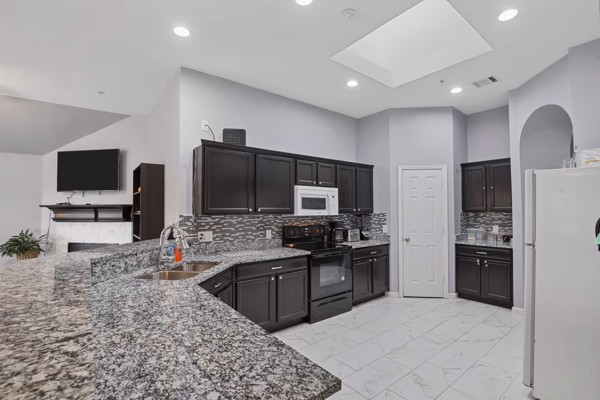 Modern kitchen with dark wood cabinets, granite countertops, a double sink, black stove, white microwave, and a white refrigerator. The kitchen has a tiled floor and a backsplash with a mosaic tile design. In the background, there is a living area with a mounted flat-screen TV and a fireplace.