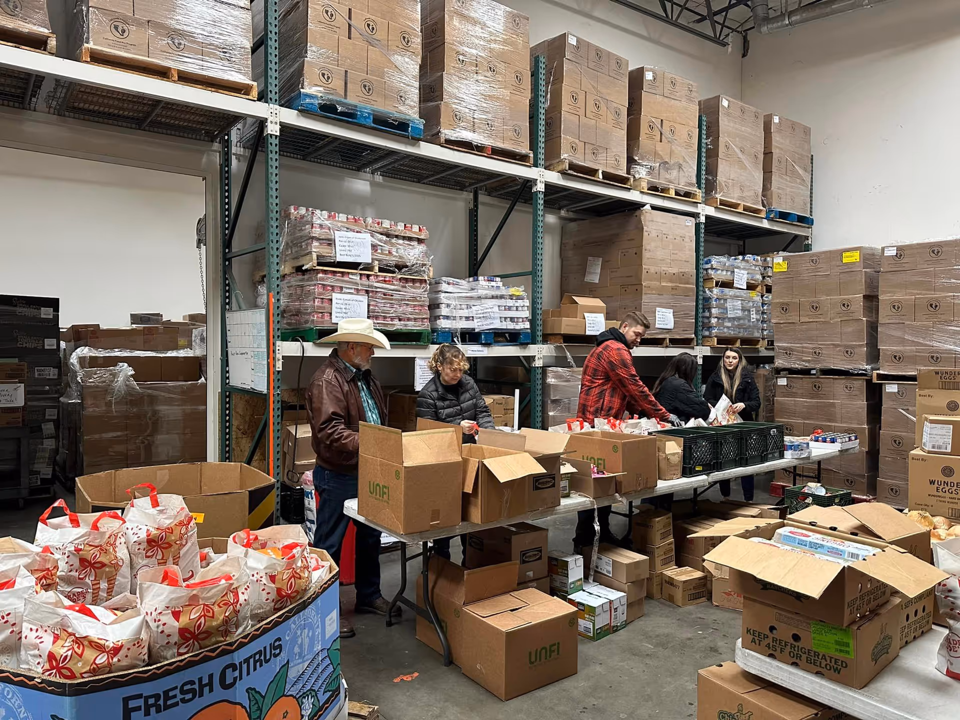 Four people working in a warehouse-like storage area with tall metal shelves stocked with large boxes and pallets. They are packing or organizing items into cardboard boxes on tables. The floor is concrete and the space is industrial with high ceilings.