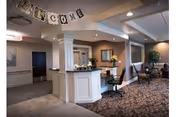 Interior view of a senior living facility reception area with a white reception desk, a chair, and a 'WELCOME' banner hanging from the ceiling. The area has carpeted flooring, recessed lighting, and seating with chairs and a small table in the background.