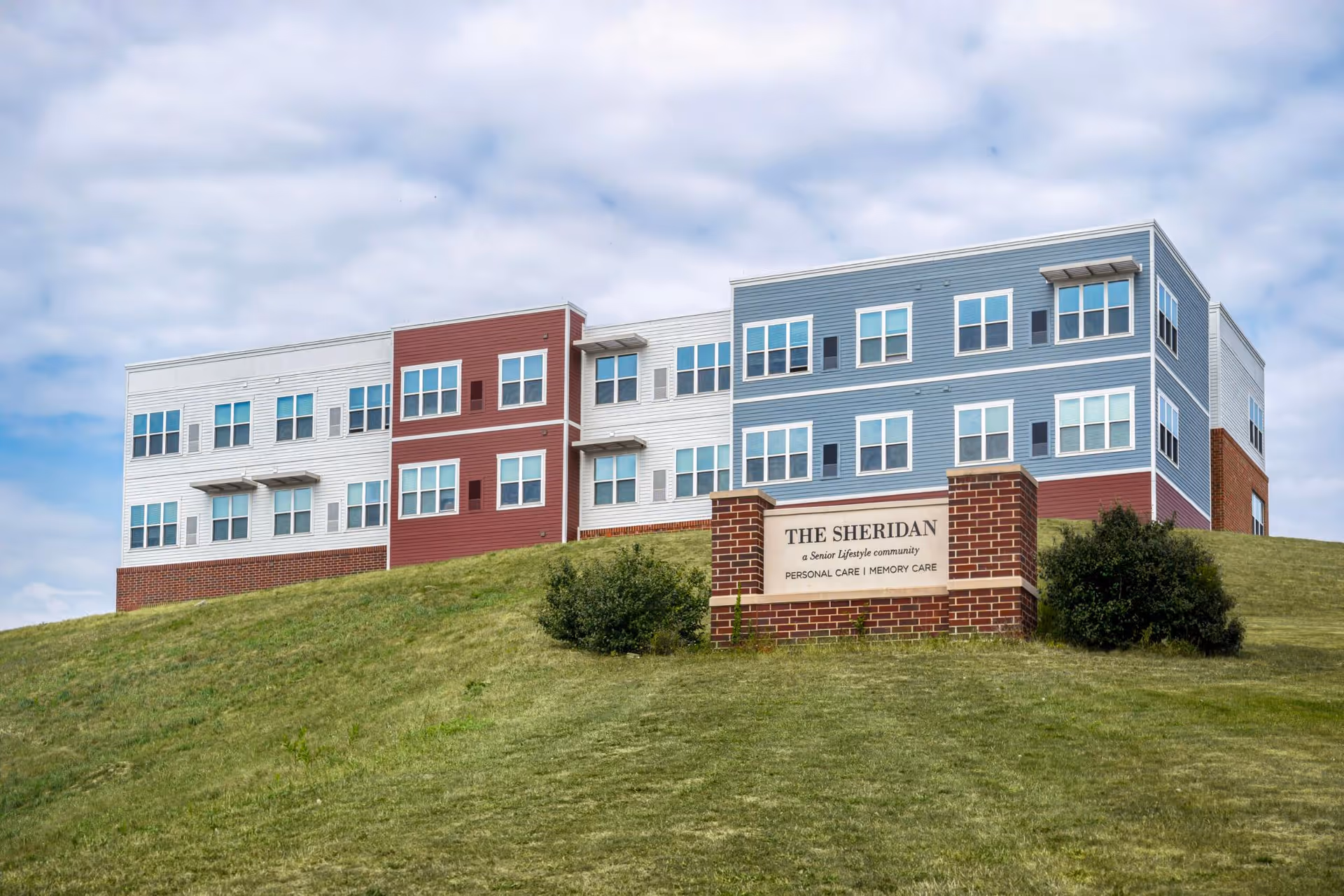 Exterior view of a senior living community building named The Sheridan, situated on a grassy hill under a partly cloudy sky. The building has multiple windows and is painted in white, red, and blue sections. A brick sign in front reads 'THE SHERIDAN a Senior Lifestyle community PERSONAL CARE | MEMORY CARE'.