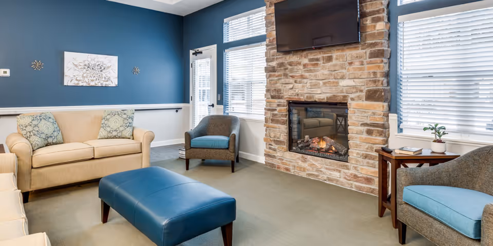 A cozy living room with a beige sofa, two gray armchairs with blue cushions, a blue leather ottoman, and a stone fireplace with a mounted flat-screen TV above it. The walls are painted blue and white, and there are windows with white blinds letting in natural light. A small wooden side table with a plant and books is beside one of the armchairs.