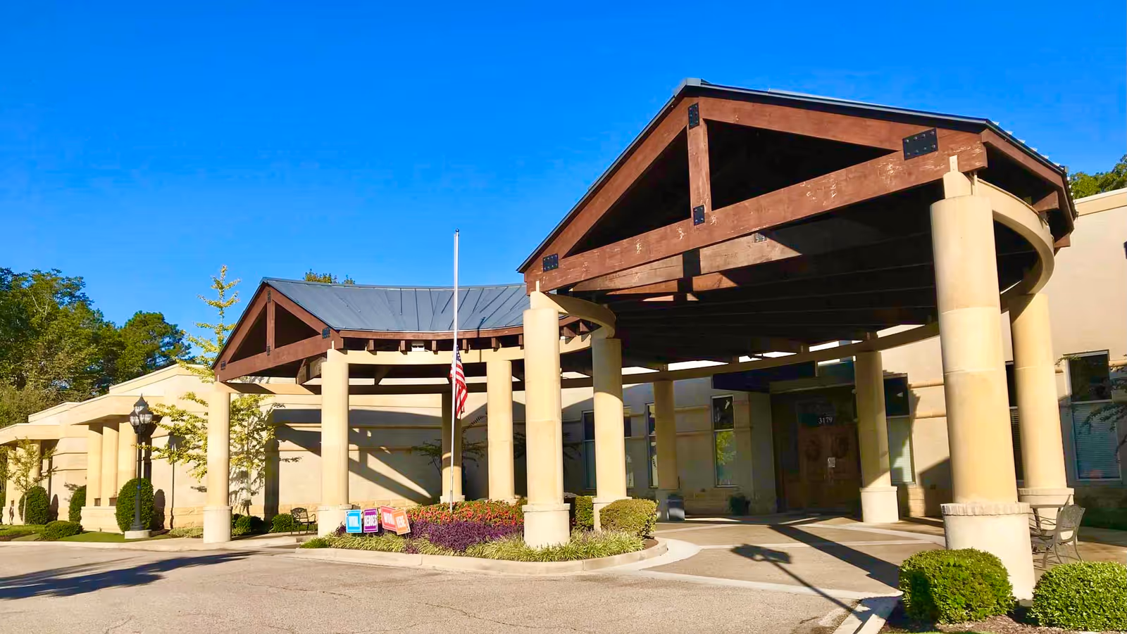 Entrance of The Gardens of Germantown facility with a covered driveway supported by large beige columns and wooden beams, an American flag at half-mast, and well-maintained landscaping under a clear blue sky.
