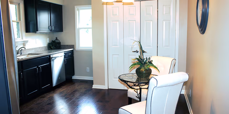 A modern kitchen area with dark wooden cabinets, a stainless steel dishwasher, and a granite countertop. There is a small round glass table with a potted plant on it and two white upholstered chairs with dark piping. The floor is dark hardwood, and there is a window letting in natural light. A round mirror hangs on the beige wall to the right.