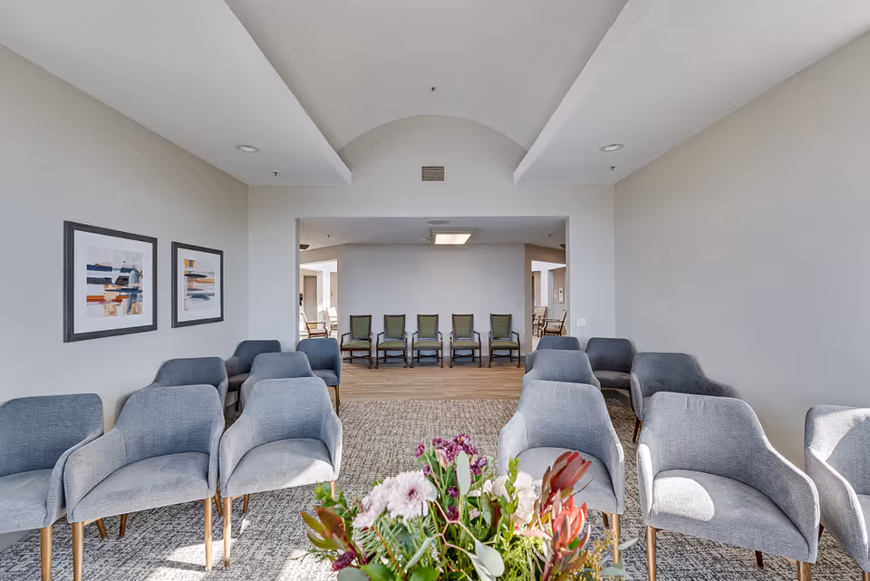 A bright and spacious seating area in a senior living facility with multiple gray upholstered armchairs arranged in rows facing a central point. There are two abstract paintings on the left wall and a bouquet of flowers in the foreground. In the background, there are additional chairs lined up against the far wall in a separate room.
