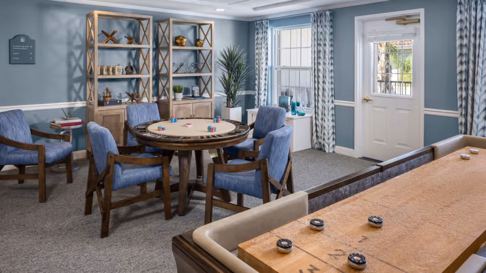 A cozy senior living common area with a round table surrounded by four blue cushioned wooden chairs, a shuffleboard table in the foreground, two wooden shelving units with decorative items, a window with patterned curtains, and a door leading outside.
