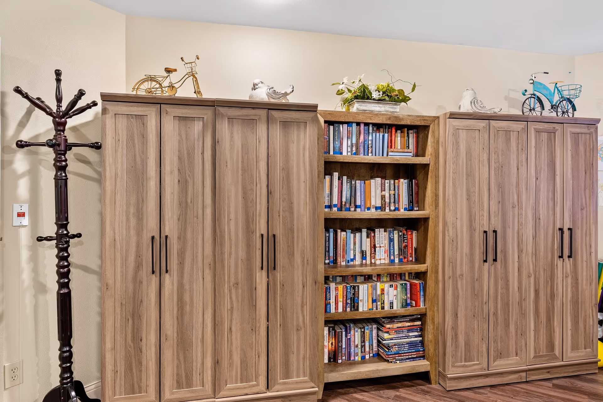 A wooden bookshelf filled with books is flanked by two wooden cabinets with closed doors. On top of the cabinets are decorative items including small bicycle models and bird figurines. To the left of the cabinets is a dark wooden coat rack standing against a beige wall.