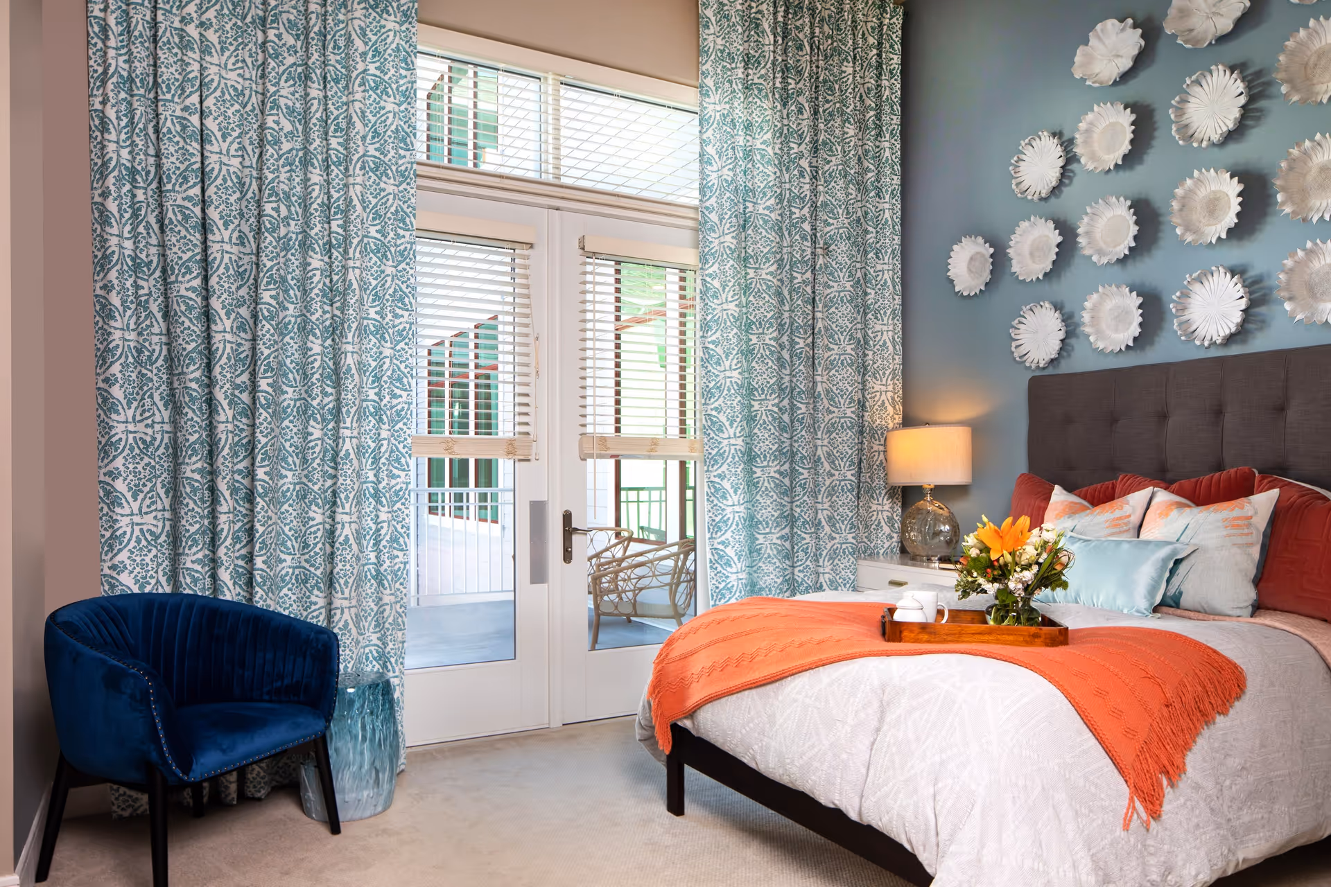 Decorated bedroom featuring a bed with an orange throw and floral pillows, teal patterned curtains over glass doors, a navy chair, and decorative wall plates on the wall.