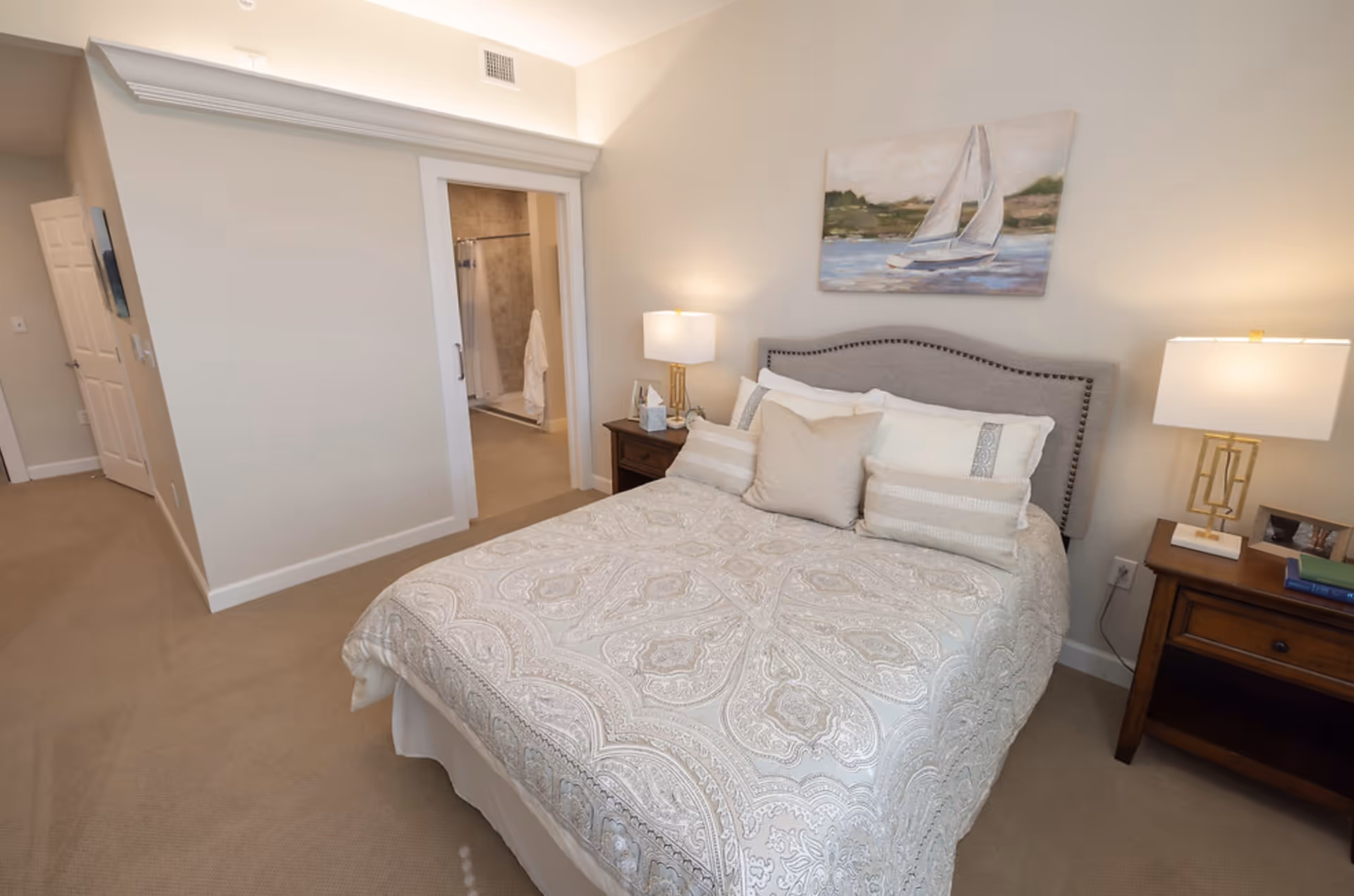 Bedroom with a patterned bedspread, upholstered headboard, two nightstands with lamps, and an open doorway to a bathroom.