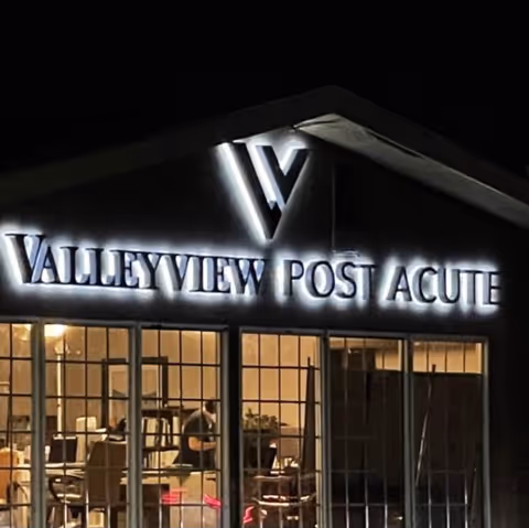 Night view of the front exterior of a building with illuminated signage reading 'Valley View Post Acute'. The building has large windows showing an interior with tables and chairs.