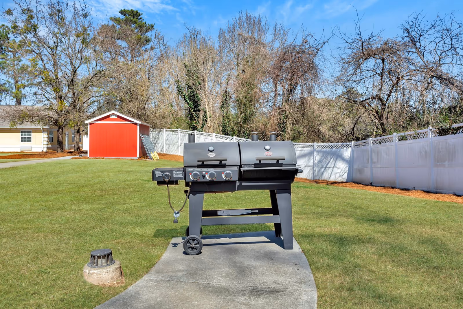 Outdoor area with a black dual-chamber grill on a concrete pad, surrounded by green grass. In the background, there is a red shed, a white fence, and leafless trees under a clear blue sky.