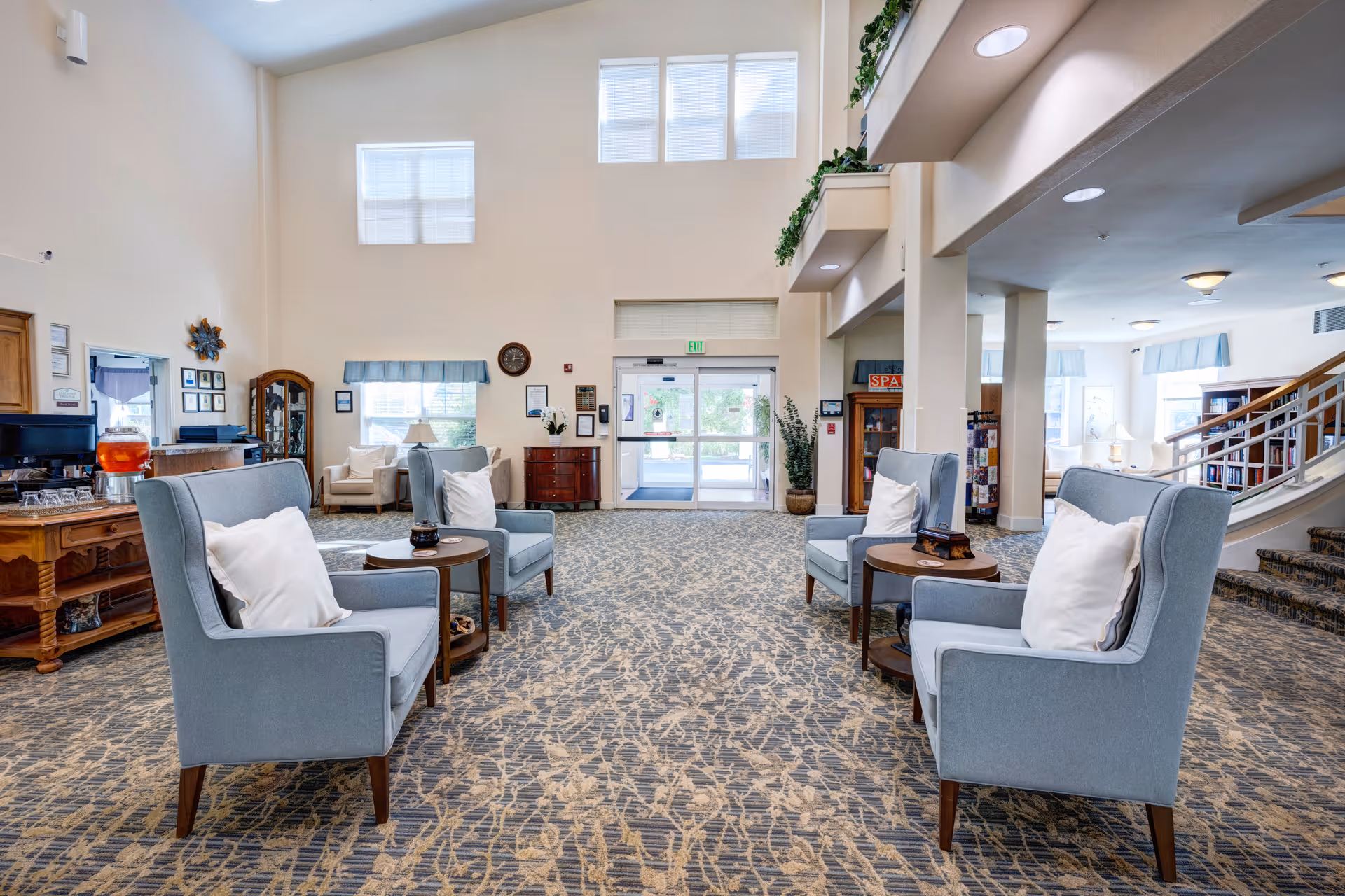 Spacious senior living lobby with high ceilings, clusters of blue armchairs and side tables facing the entrance doors.