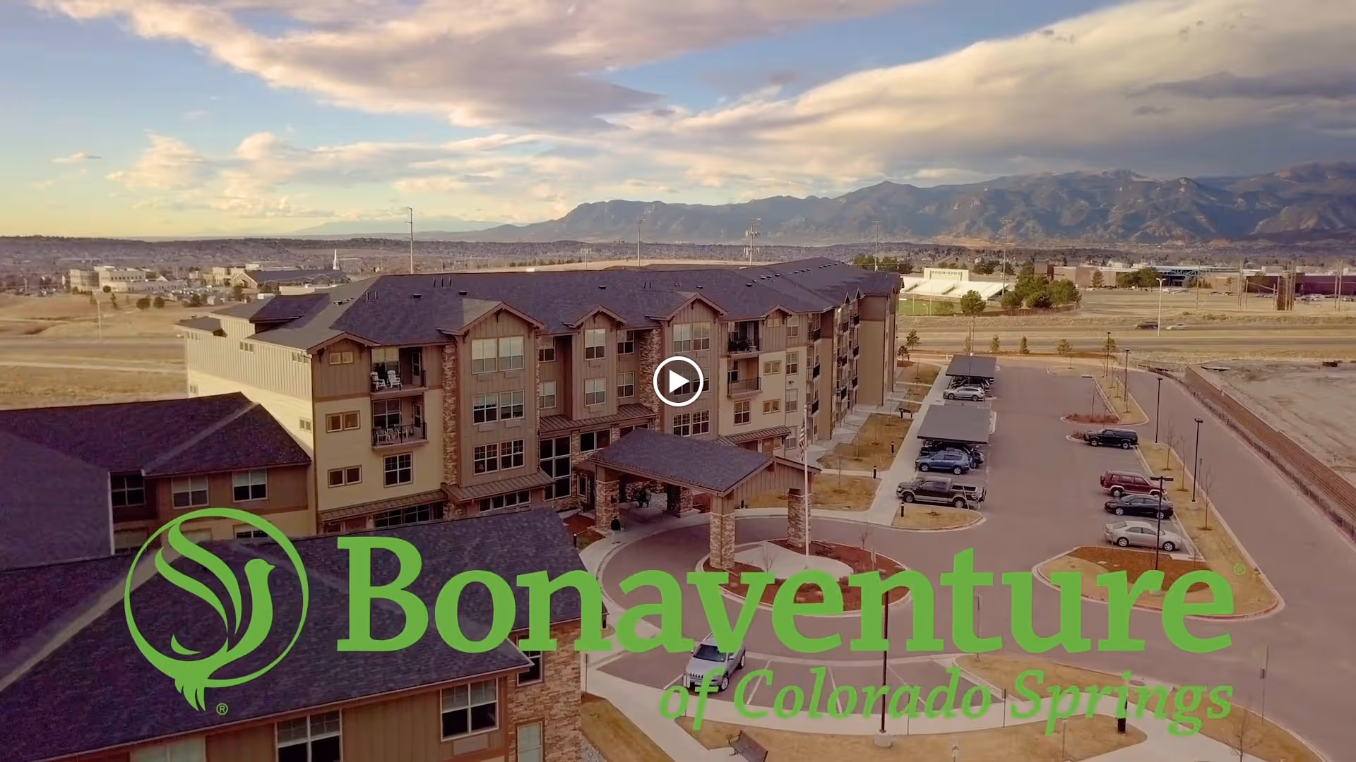 Aerial view of a multi-story senior living facility building with a parking lot and surrounding landscape, set against a backdrop of mountains and a partly cloudy sky.