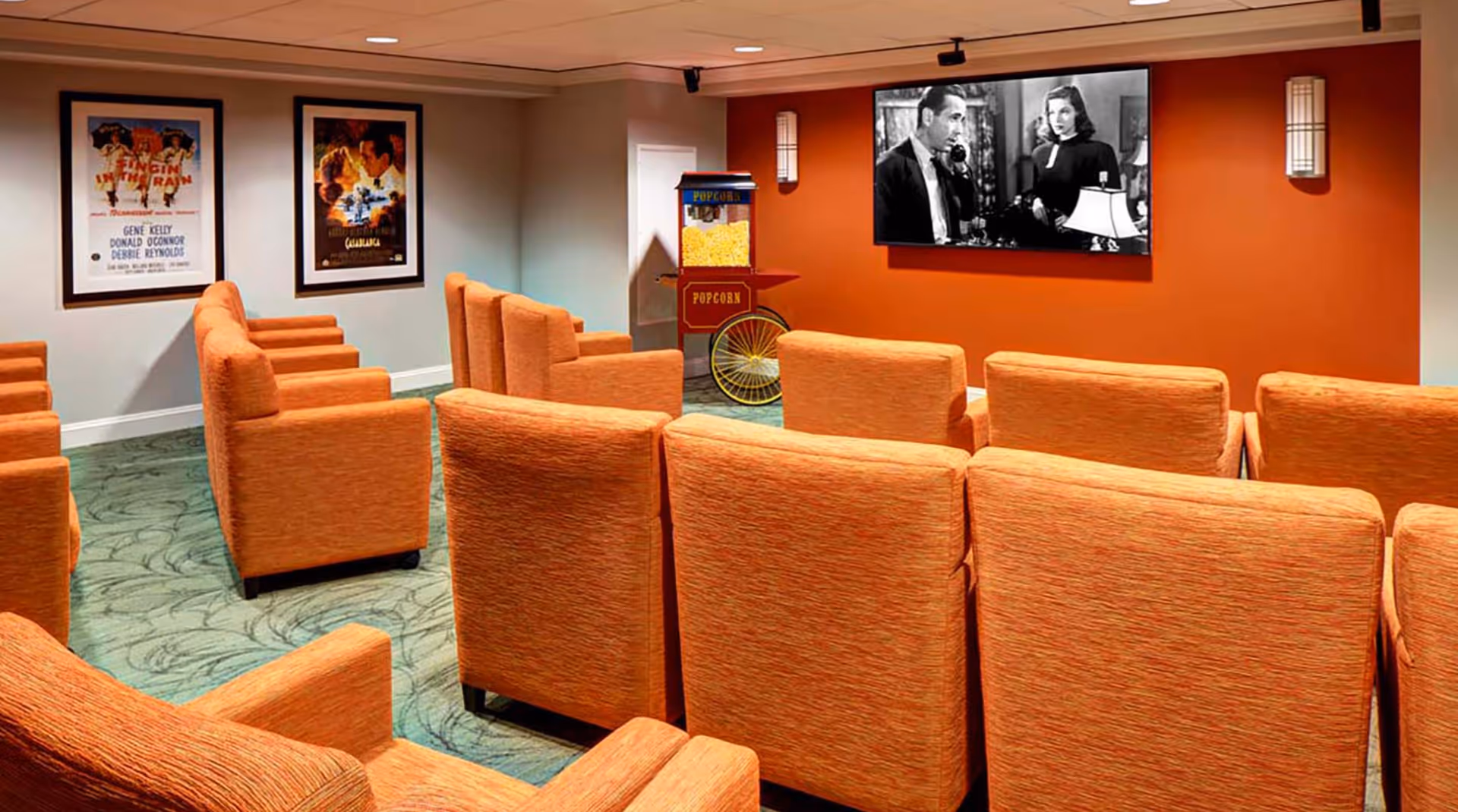 A small movie theater room with rows of orange upholstered chairs facing a large screen showing a black-and-white movie. There is a vintage-style popcorn machine near the screen, and two framed classic movie posters on the wall to the left. The walls are painted in light gray and orange tones.