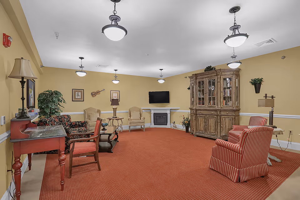 A cozy living room with yellow walls and red carpet. The room features various seating options including two striped armchairs, two beige armchairs, and a patterned loveseat. There is a wooden cabinet with glass doors, a wall-mounted TV above a white fireplace, several lamps, framed pictures, and decorative plants.