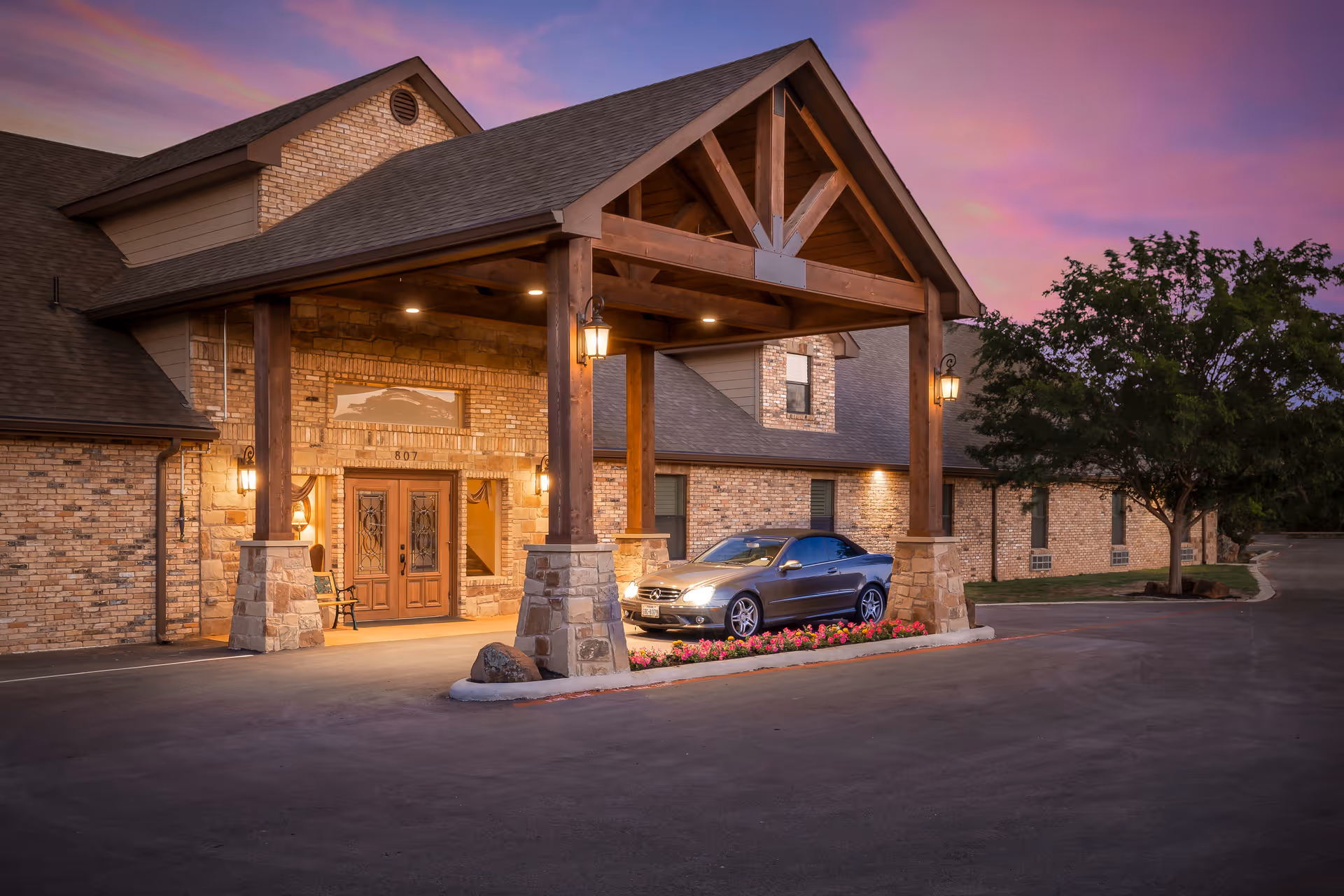 Exterior view of a brick building with a covered entrance supported by wooden beams and stone pillars. A silver convertible car is parked under the entrance canopy, and the sky is a colorful sunset with shades of purple and pink. There are some trees and flowers near the entrance.
