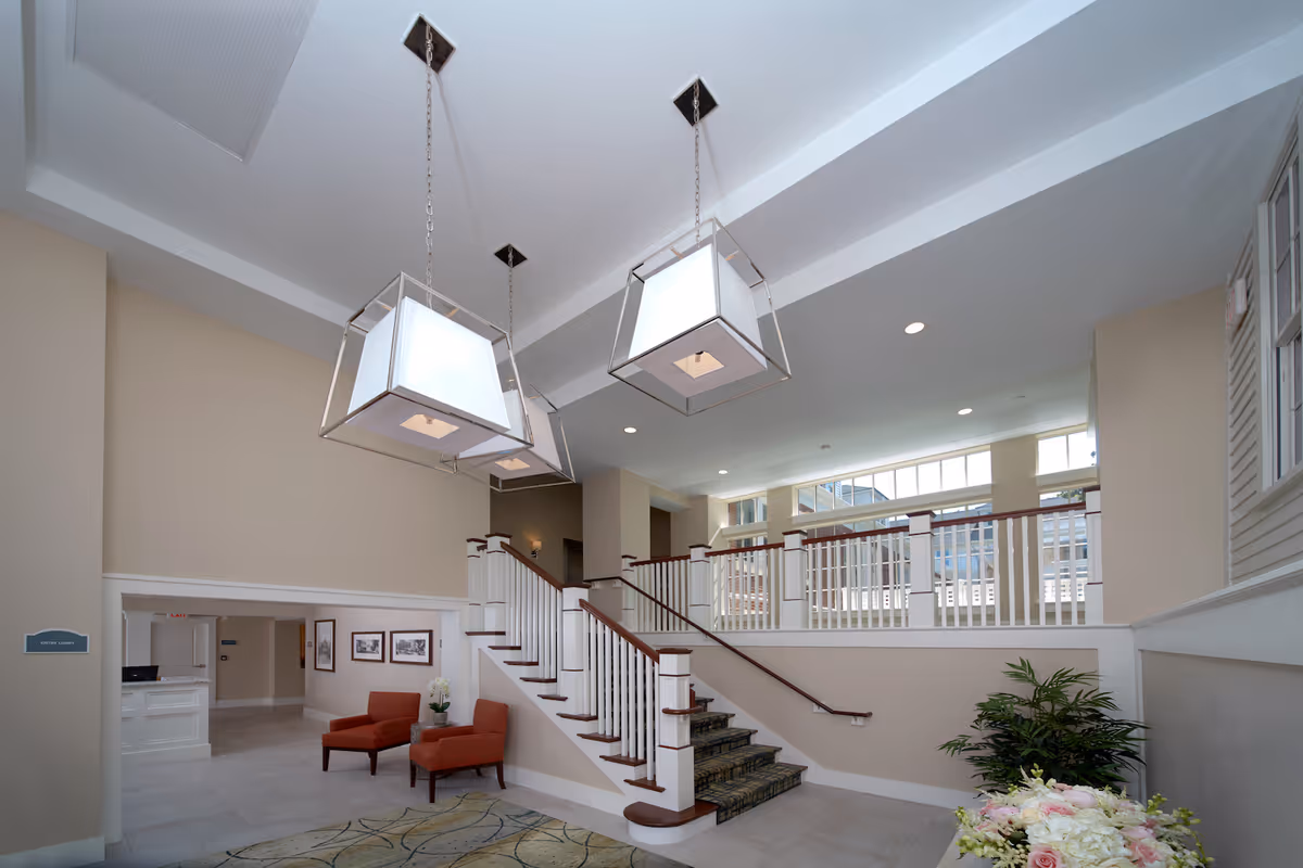 Bright senior living lobby with a central staircase, modern hanging lights, seating area and plants.