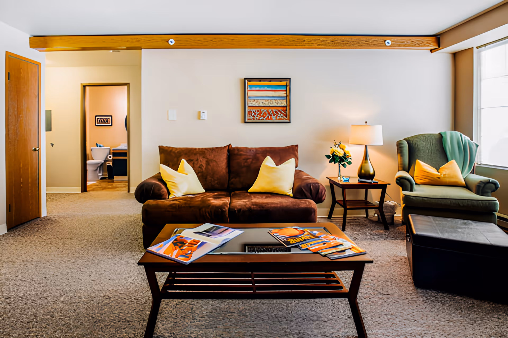 A cozy living room with a brown sofa adorned with two yellow pillows, a wooden coffee table with magazines on it, a green armchair with a yellow pillow and a green throw blanket, a side table with a lamp and a vase of yellow flowers, and a window letting in natural light. In the background, an open doorway reveals a bathroom with a toilet and a framed picture on the wall.