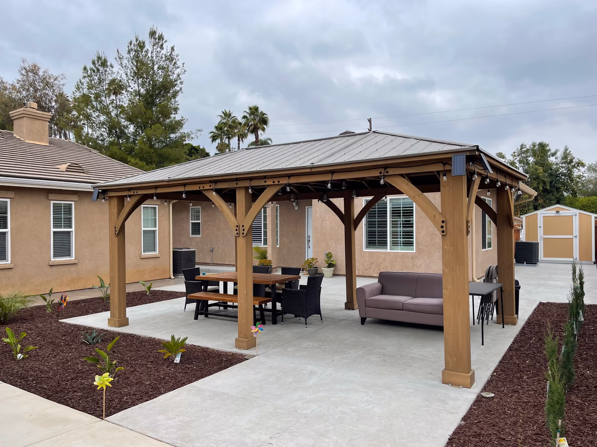 Outdoor patio area with a wooden pergola featuring string lights. Under the pergola, there is a wooden picnic table with benches and black chairs, as well as a gray sofa and a small black table. The patio is surrounded by mulched garden beds with small plants and flowers. In the background, there are beige buildings with windows and a shed. The sky is cloudy.
