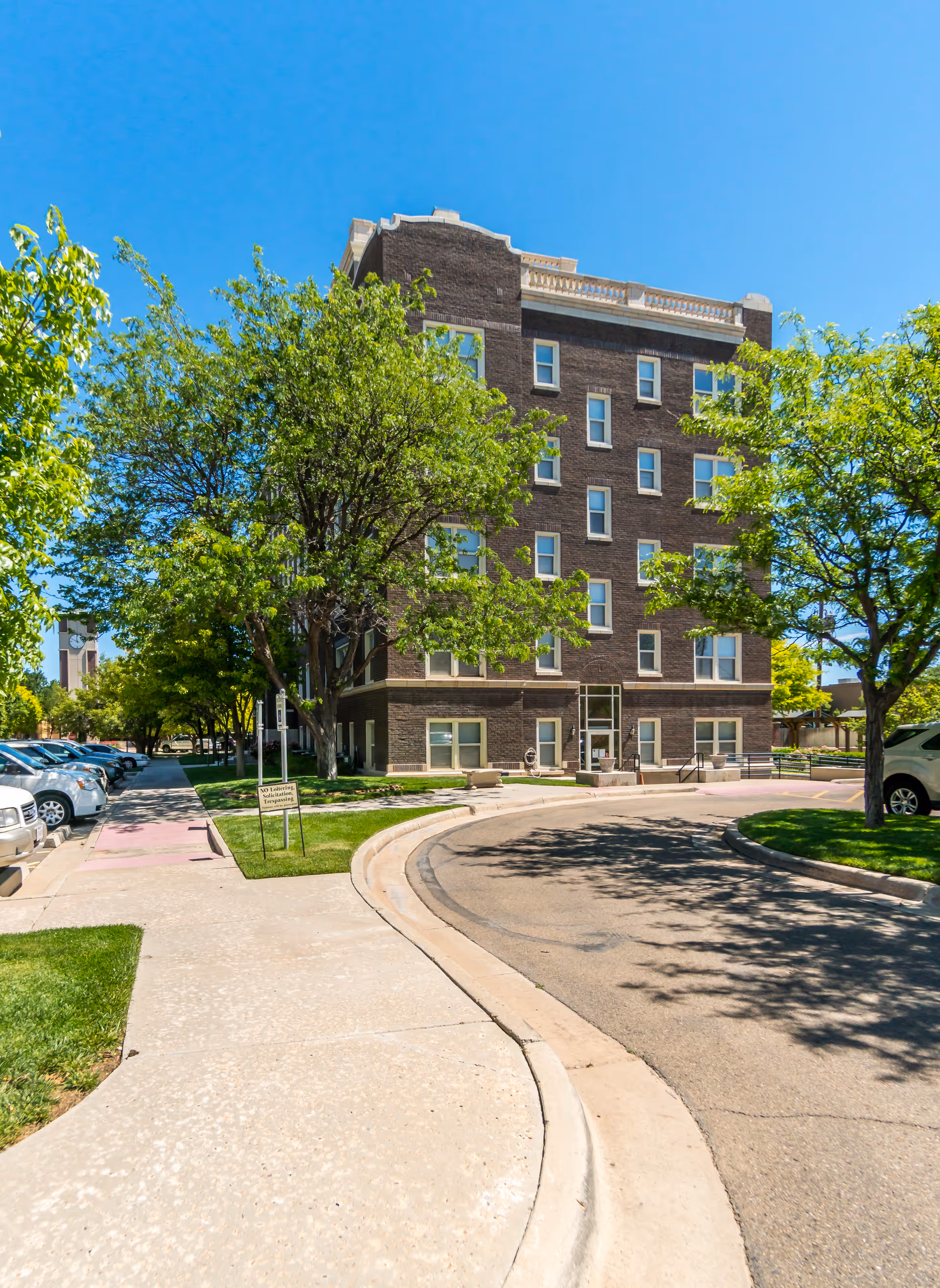 Exterior view of a multi-story brick building with several windows, surrounded by green trees and a clear blue sky. There is a curved driveway and a sidewalk with parked cars along the side.
