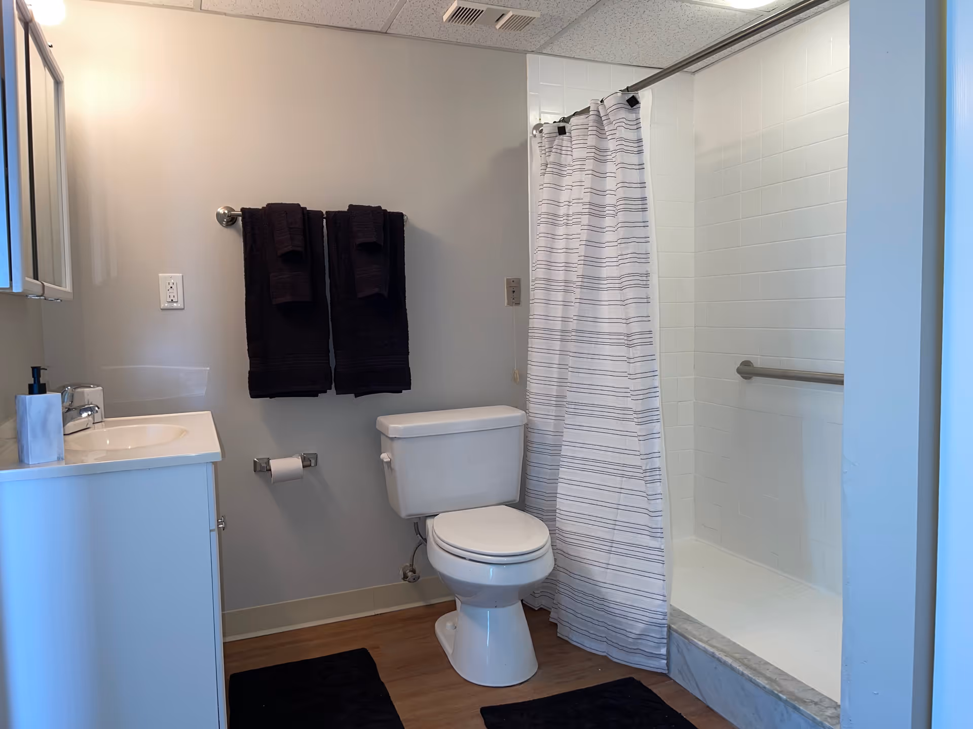 A clean bathroom featuring a white toilet, a white sink with a soap dispenser, two dark towels hanging on a towel rack, a toilet paper holder with a roll of toilet paper, and a shower area with a white and gray striped shower curtain and a grab bar inside the shower.