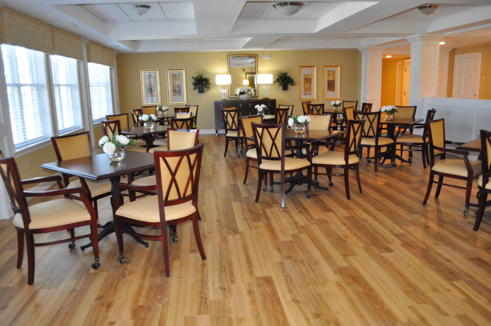 A bright and spacious dining room with multiple wooden tables and chairs arranged neatly. Each table has a small vase with white flowers. The room features large windows with blinds on the left side, wooden flooring, and soft yellow walls decorated with framed artwork and plants. There is a sideboard with lamps and a mirror on the far wall.