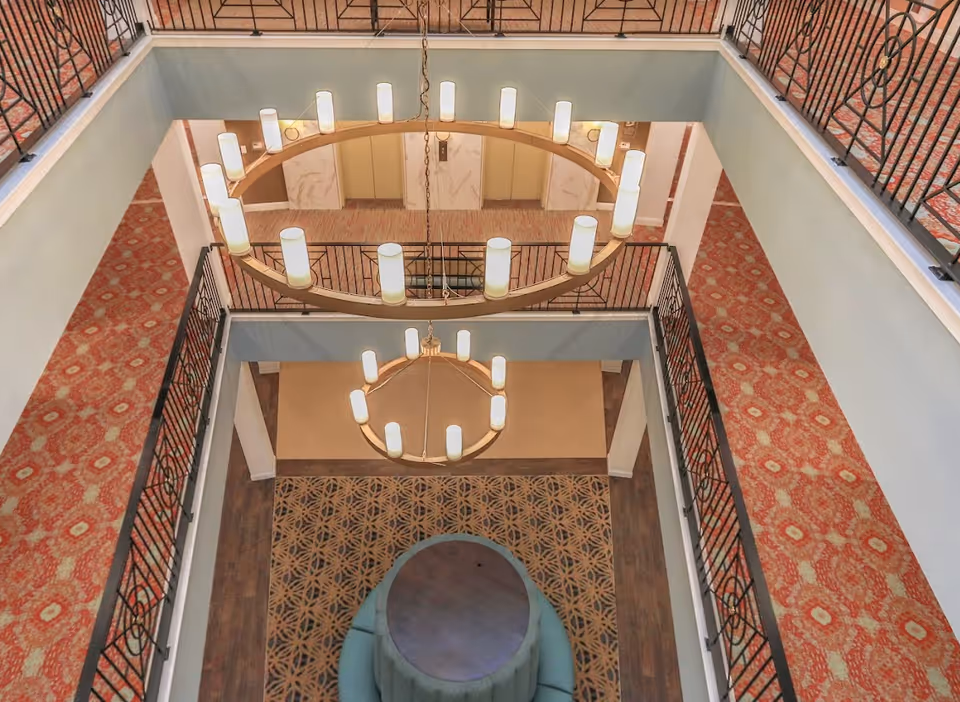 View looking down into a multi-level interior space with two circular chandeliers hanging from the ceiling. The area features patterned carpets, a round cushioned seating area on the lower level, and elevator doors on the far side. Black metal railings surround the upper levels.