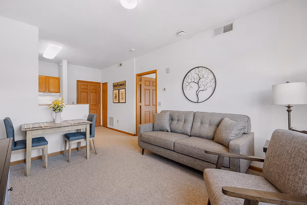 Bright open-plan living room with a gray sofa and armchair, small dining table, and wall art.