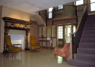 Interior view of a spacious living area with a large ornate fireplace, two green armchairs, a red upholstered chair, a wooden table with decorative items, and a staircase with a dark railing leading to an upper floor with a balcony.