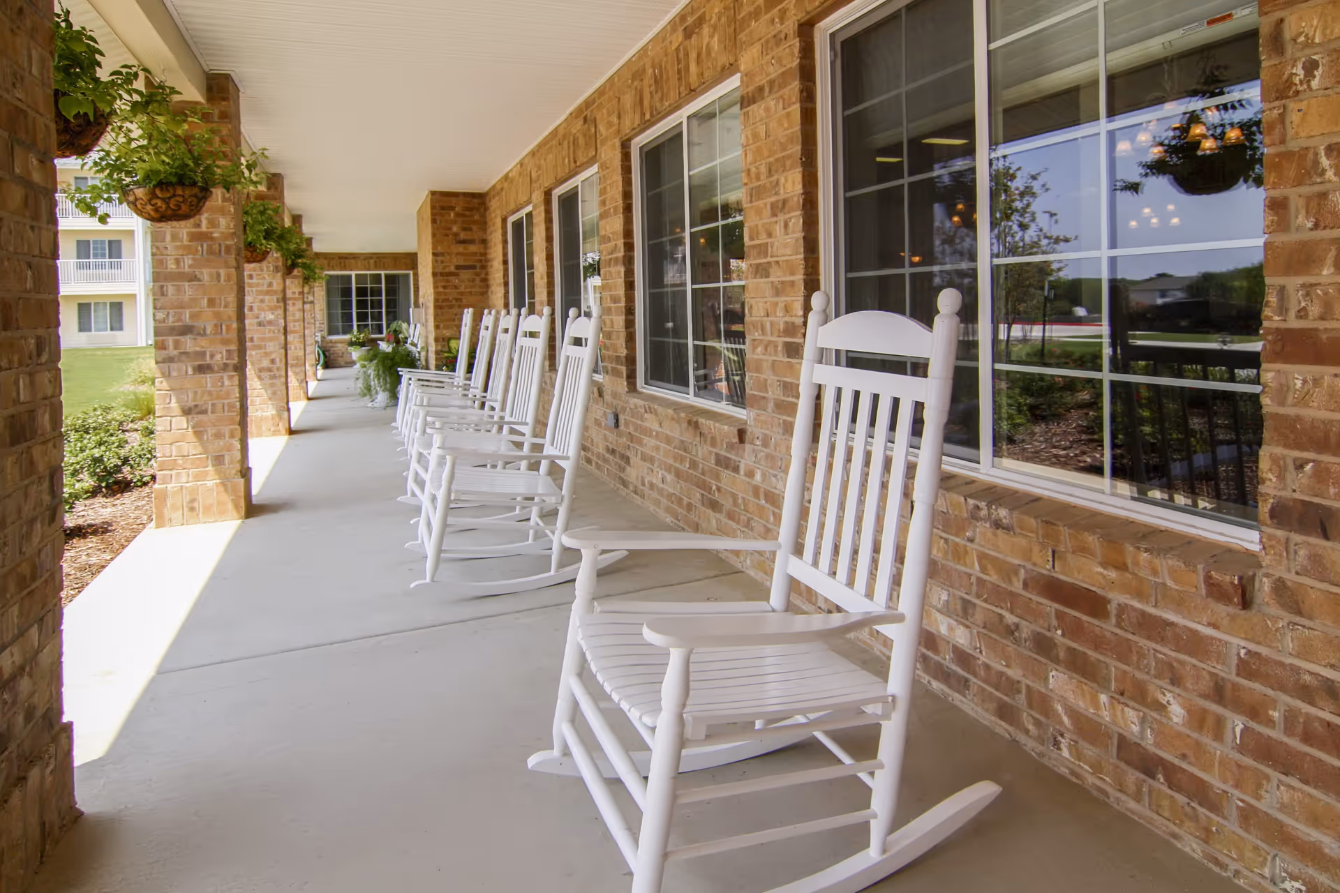 A covered outdoor porch area with a row of white wooden rocking chairs lined up against a brick wall with windows. Hanging plants are suspended from the ceiling, and greenery is visible outside.