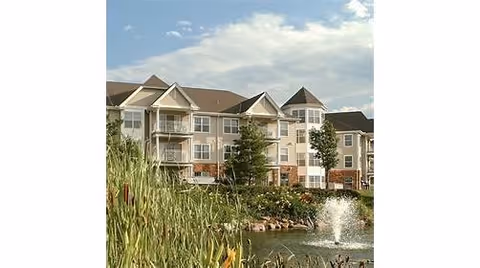 Exterior view of a multi-story senior living facility named Immanuel: Lakeside, featuring beige and white siding with balconies, surrounded by greenery and a pond with a water fountain in the foreground under a partly cloudy sky.