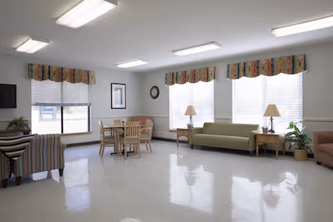 Bright communal lounge with sofas, chairs, a small dining table, windows with valances, lamps, and overhead lighting.