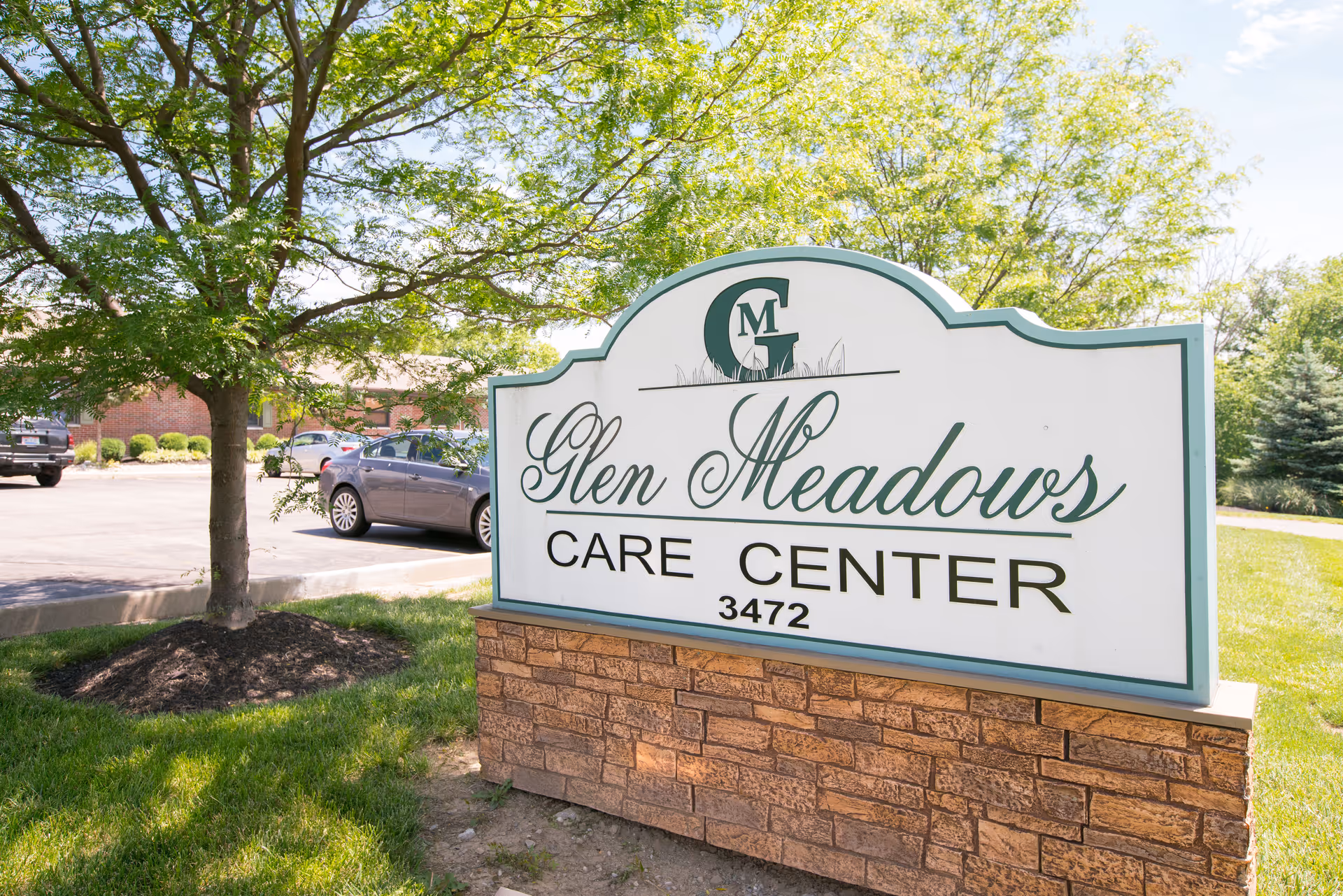 Large outdoor sign that reads "Glen Meadows Care Center 3472" beside a tree and parking lot.
