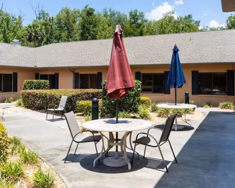 Outdoor courtyard area with round tables and chairs. Each table has a closed umbrella, one red and one blue. The courtyard is surrounded by a single-story building with windows and a shingled roof. There are bushes and greenery along the edges of the paved area under a partly cloudy sky.