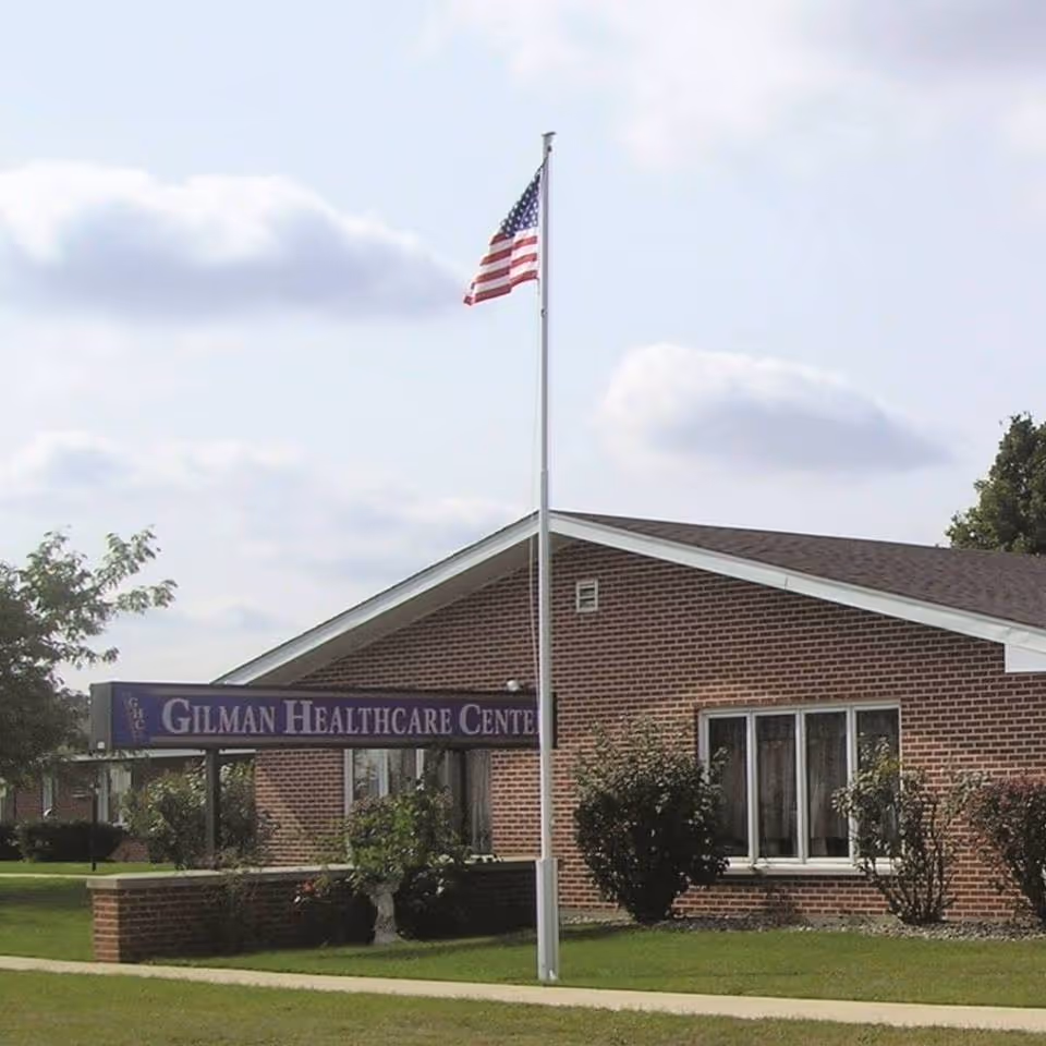 Brick single-story healthcare building with an American flag on a flagpole and a sign reading "Gilman Healthcare Center" in front.