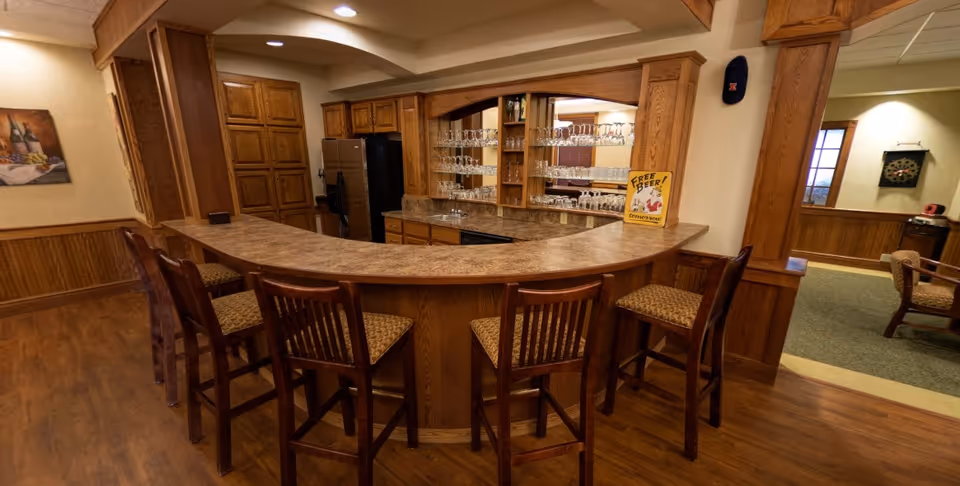 Interior view of a wooden bar area with a curved countertop and six high chairs with patterned cushions. Behind the bar are shelves filled with glassware, a refrigerator, and wooden cabinets. The room has wood-paneled walls and a hardwood floor, with a small sign on the bar that reads 'Free Beer Tomorrow'.