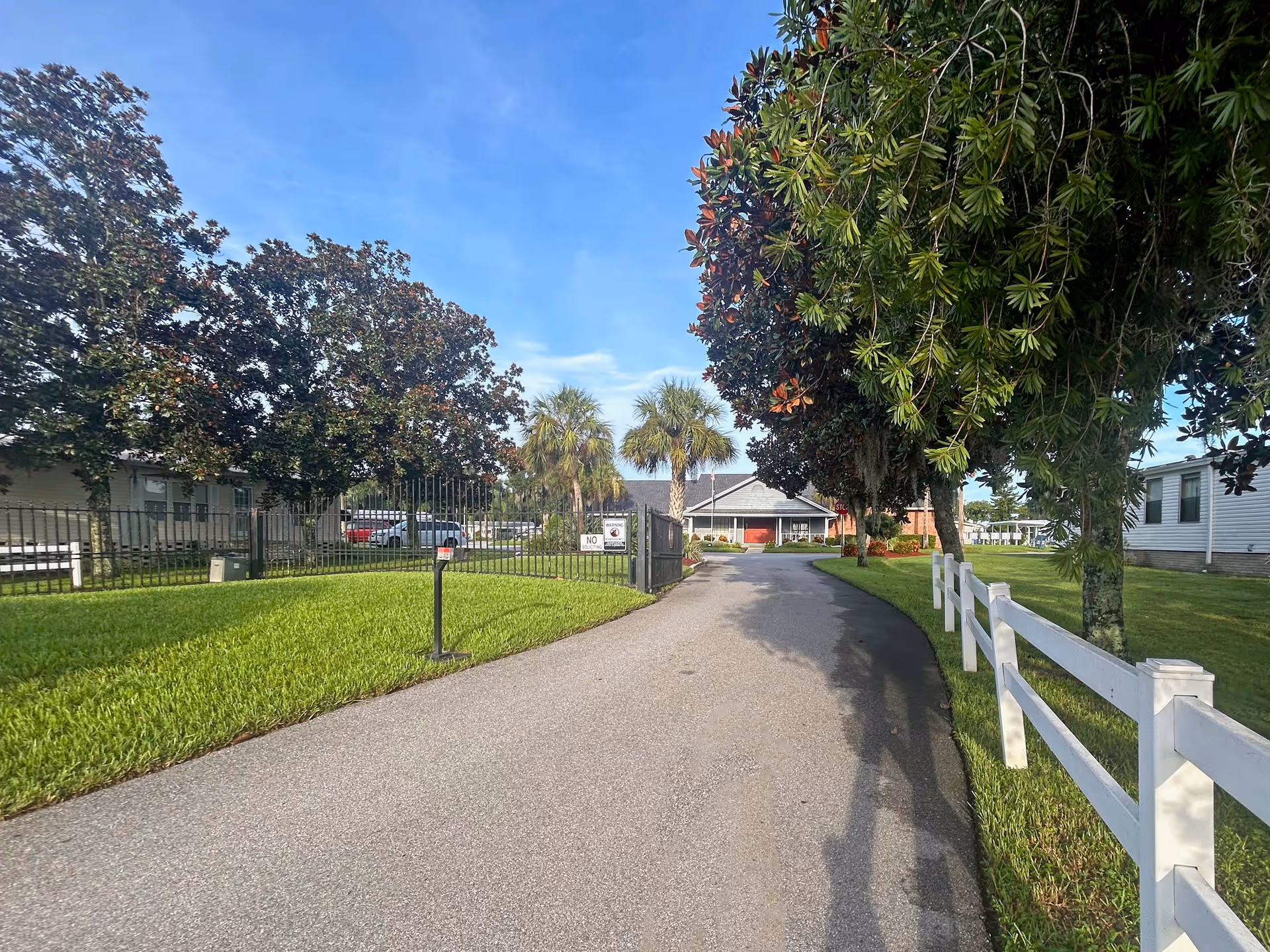 A paved driveway lined by a white fence, grass and trees leading to the entrance of a single-story retirement community building.
