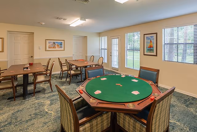 A well-lit room with multiple tables and chairs, including a round poker table with playing cards and poker chips, and rectangular tables in the background. The room has beige walls, carpeted floors, framed artwork on the walls, and windows with blinds letting in natural light.