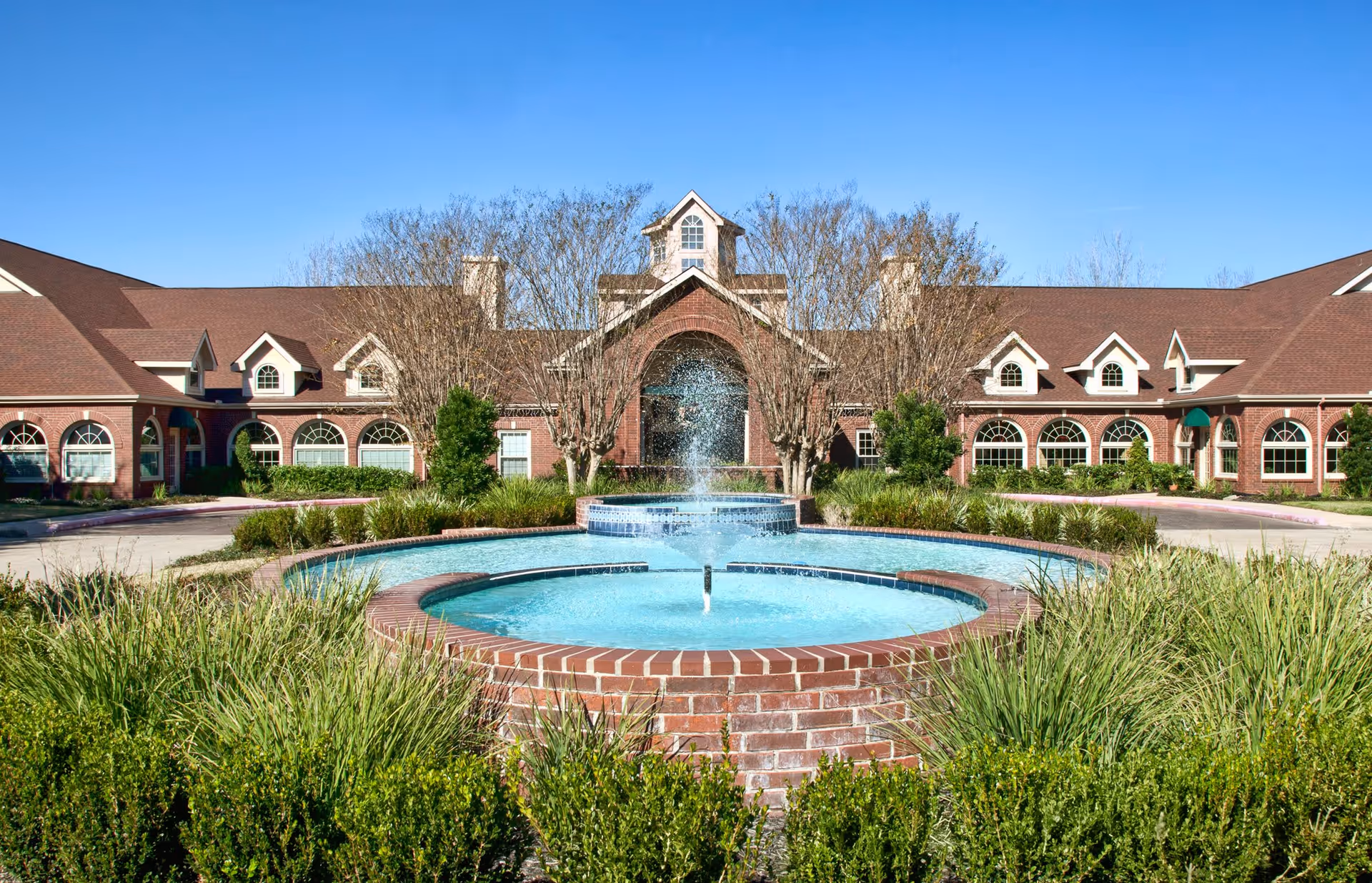Front exterior view of a senior living facility named Village on the Park Friendswood with a large circular brick fountain in the foreground, surrounded by green bushes and grass, and a clear blue sky above.