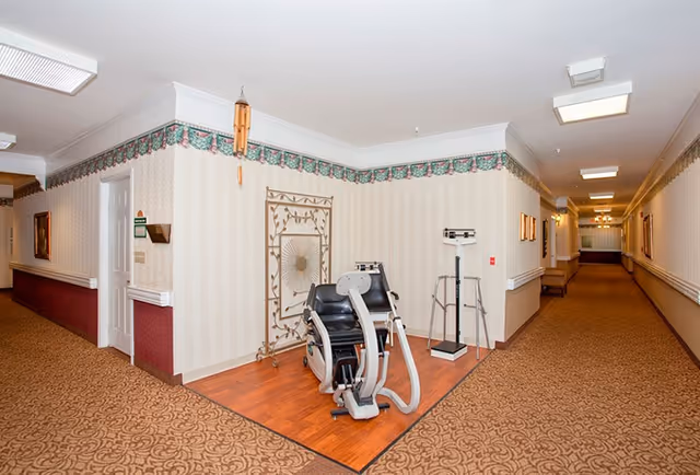 Carpeted senior living hallway with a small exercise/medical station including a scale and a reclining exercise chair on a wood floor inset.