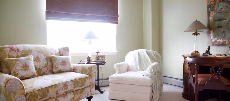 A cozy living room area with a floral patterned sofa, a white armchair draped with a light blanket, a small round side table with a lamp and picture frame, and a wooden desk with a lamp and chair. A window with a brown roman shade is in the background.