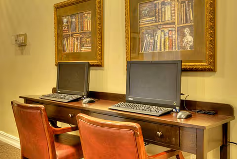 Two desktop computers sit on a wooden desk with two leather chairs beneath framed bookshelf paintings on the wall.