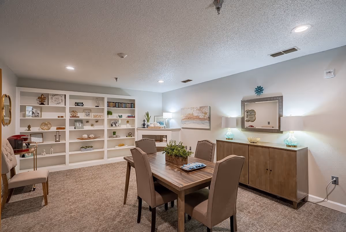 A well-lit interior room with a wooden dining table surrounded by four upholstered chairs. Behind the table is a wooden sideboard with two lamps and a decorative mirror above it. On the left side of the room, there is a large white built-in bookshelf filled with various decorative items and books. The room has carpeted flooring and neutral-colored walls.