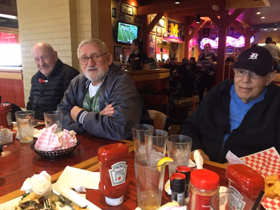 Three elderly men sit together at a restaurant table with glasses, ketchup bottles, and plates while a bar and neon signs are visible behind them.